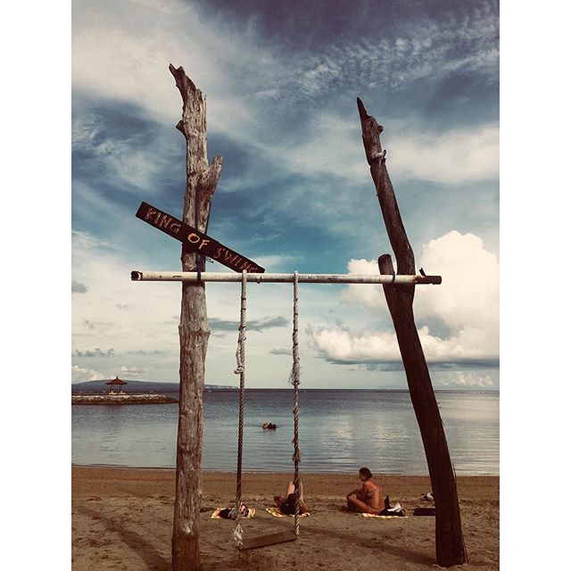 A swing set on a beach with a couple relaxing in front of the ocean.