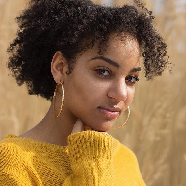 A woman with curly hair and hoop earrings gazes thoughtfully into the camera.