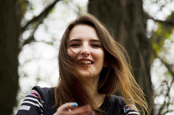 A smiling young woman with brown hair enjoys a sunny day outdoors in a park setting.