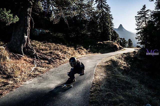 A skateboarder is riding down a winding mountain road with trees and mountain views.