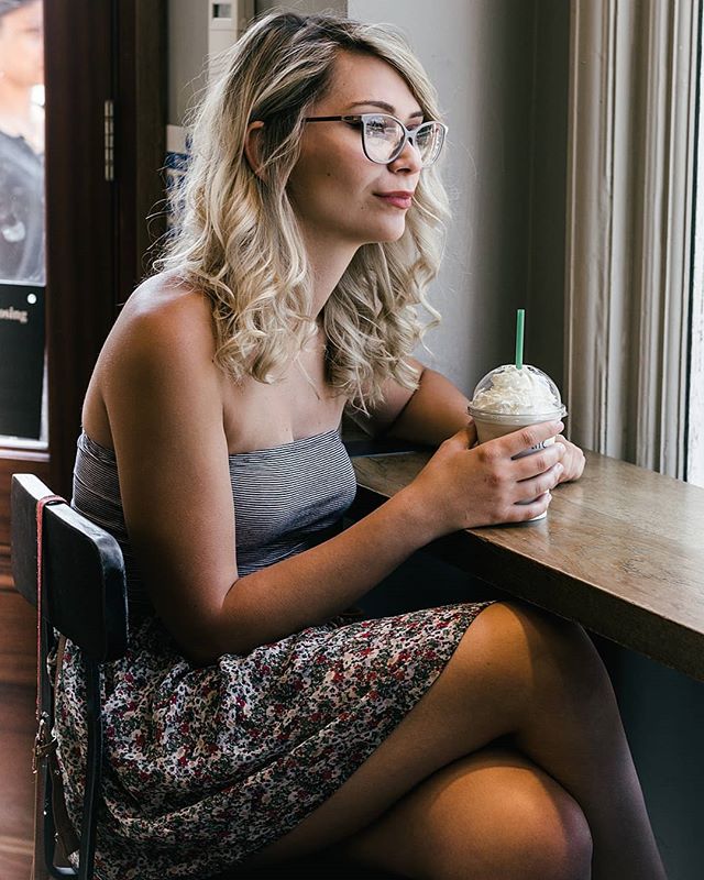 A woman sits by a window in a cafe, enjoying a coffee drink in a striped top and floral dress, looking thoughtful.