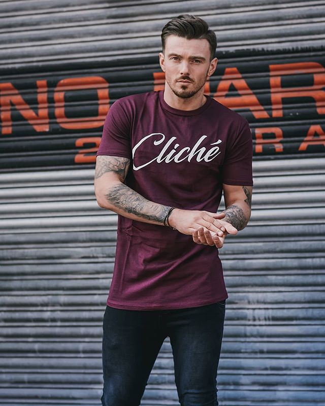 A confident man poses in a maroon t-shirt and black jeans in an urban setting.