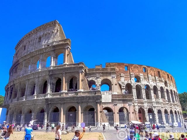 The Colosseum in Rome, Italy, stands against a clear blue sky, with many people visiting the historical landmark.