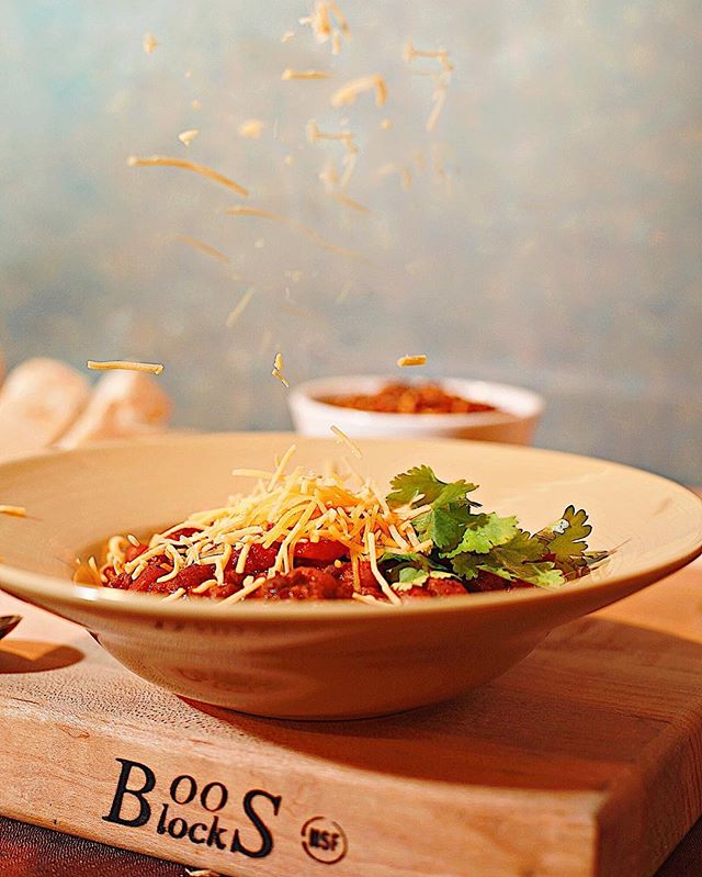 A bowl of chili is topped with cheese and cilantro on a Boos Block cutting board.