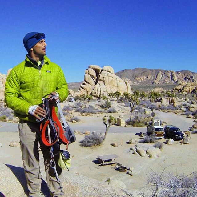 Man holding climbing gear in a desert landscape with rock formations under a clear blue sky.