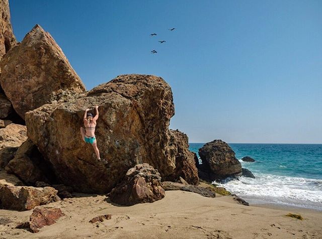 A woman rock climbs on a boulder at the beach on a clear day by the ocean.