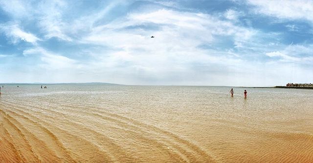 Two people are walking in the sea at a beach on a sunny day with blue skies.