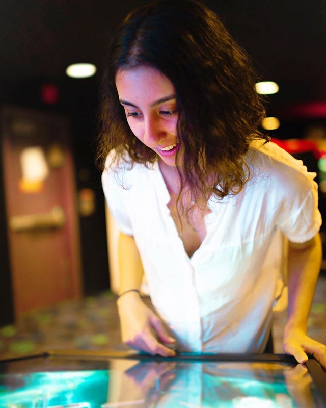 A smiling woman in a white shirt interacts with a touchscreen display in a dimly lit arcade.