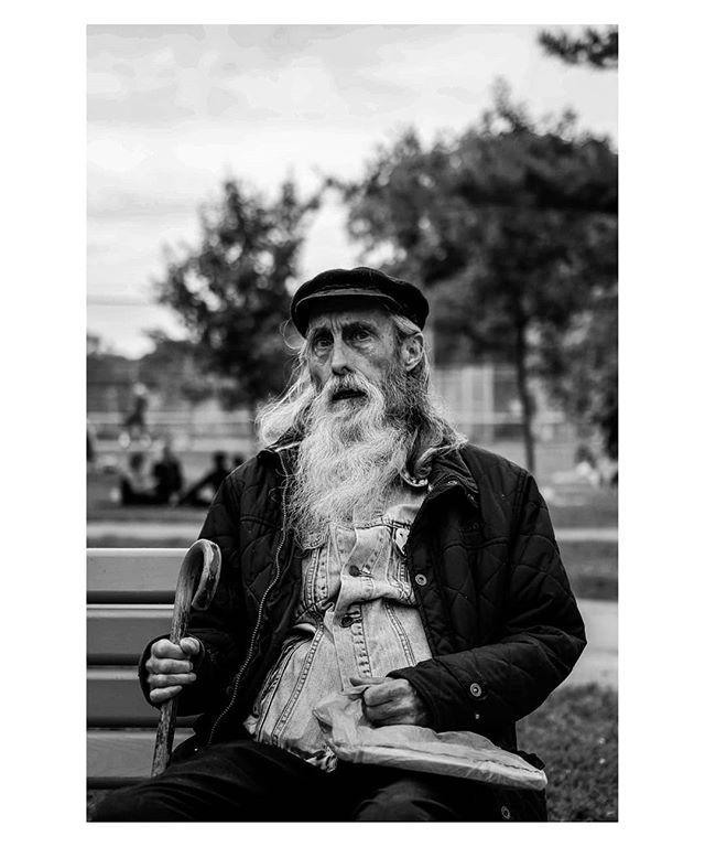 A thoughtful elderly man with a long beard sits on a park bench in a black and white portrait.