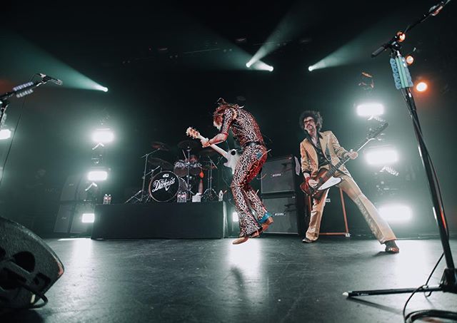 A rock band performs live on stage with dynamic lighting and energetic musicians in a wide shot.