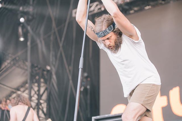 A bearded singer with a bandana performs on stage at an outdoor concert.