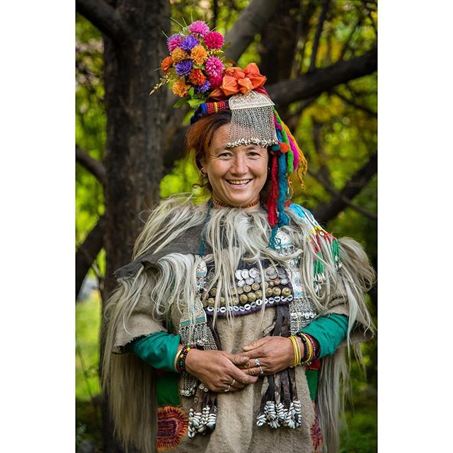 A smiling woman in traditional attire with a colorful headdress poses outdoors in front of a tree.