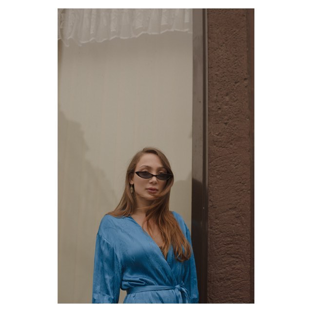 A stylish woman in sunglasses and a blue dress poses outdoors with an urban backdrop.