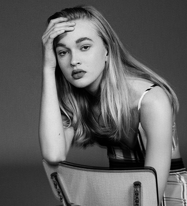 A blonde woman poses in a monochrome studio portrait, her expression serious and contemplative.