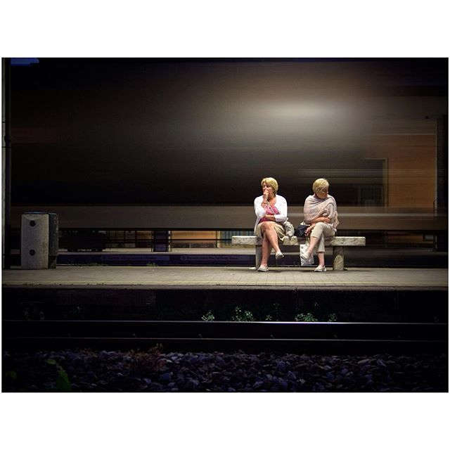 Two women sit on a bench at a train station platform, waiting with a blurred train passing by.