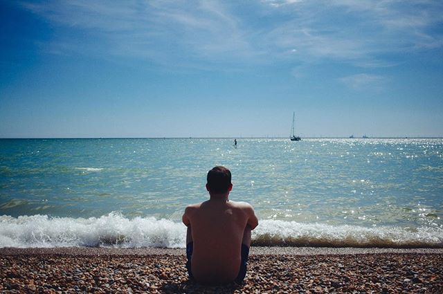 A shirtless man sits on a pebble beach, gazing out at the ocean and the distant horizon under a clear, blue sky.