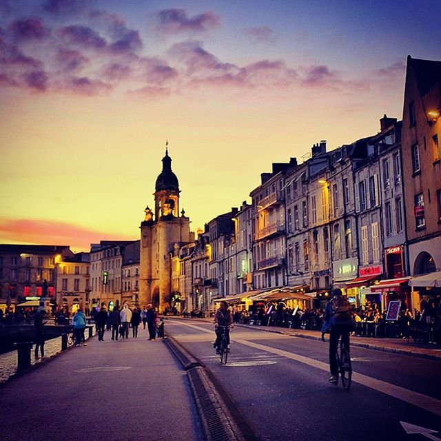 A picturesque city street scene at sunset with people walking and cycling along the road.