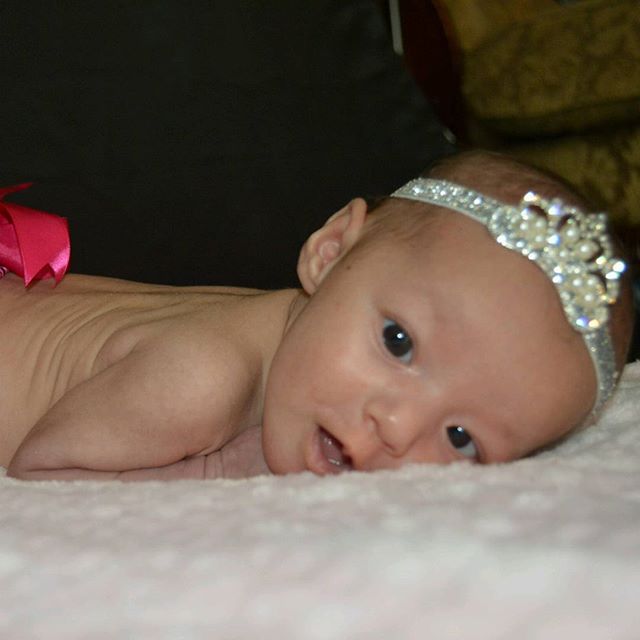 A sweet newborn baby girl lies on a soft, pink blanket with a sparkly headband.