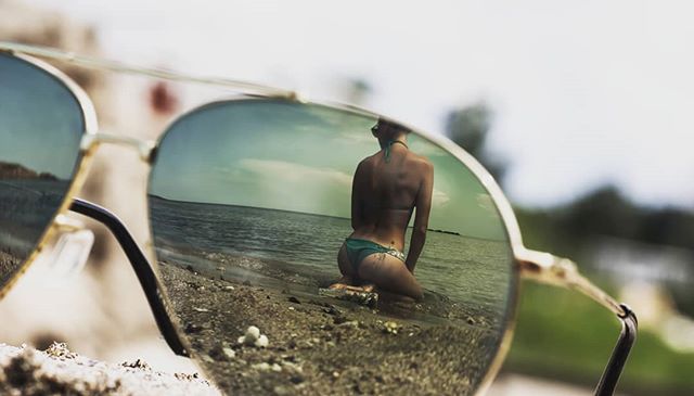A woman is reflected in sunglasses, sitting on a sandy beach near the ocean on a sunny day.