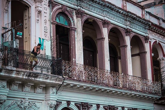 A woman stands on a balcony of an old building in a city, looking out at the street.