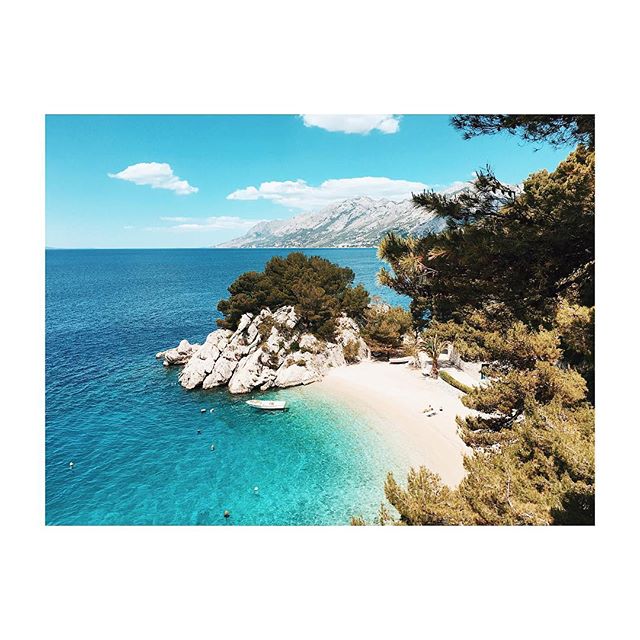 A scenic coastal view showcases a turquoise sea meeting a white sand beach against a backdrop of mountains and blue skies.