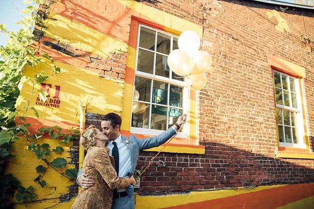 A happy couple share a kiss while holding balloons in front of a colorful building. 