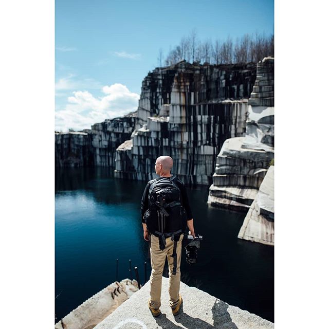 A man with a backpack stands on a cliff overlooking a scenic lake in an abandoned quarry.