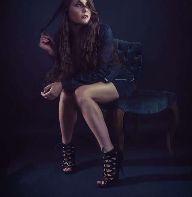 A woman sits in a chair wearing heels and a short skirt, in a moody, editorial style.