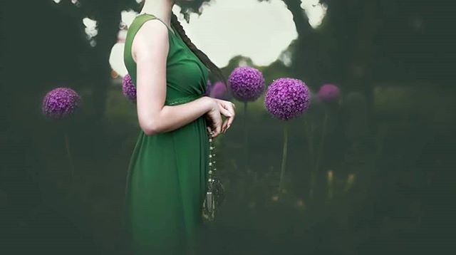 A woman in a green dress stands among dreamy purple flowers in a tranquil garden setting.
