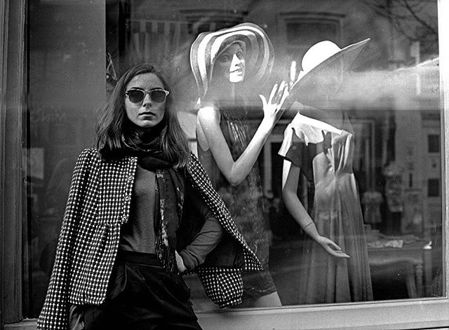A woman stands in front of a shop window with mannequins, capturing a vintage street style moment.