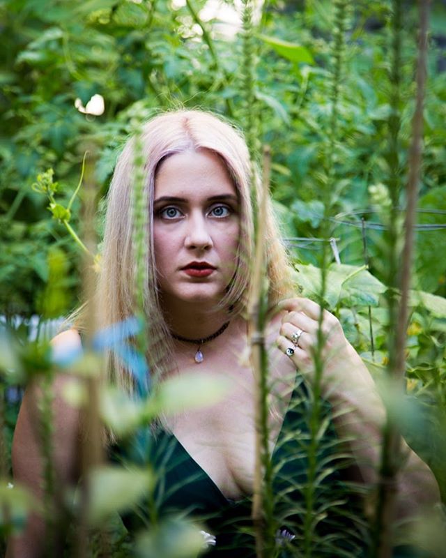 A woman with blond hair poses outdoors surrounded by lush green foliage, creating a dreamy and mysterious portrait.