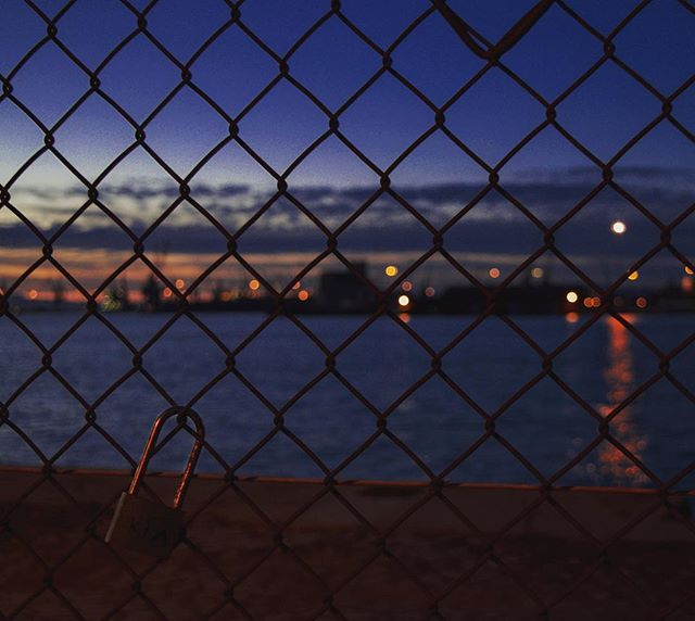 A padlock hangs on a chain-link fence at night, with blurred city lights and water in the background.