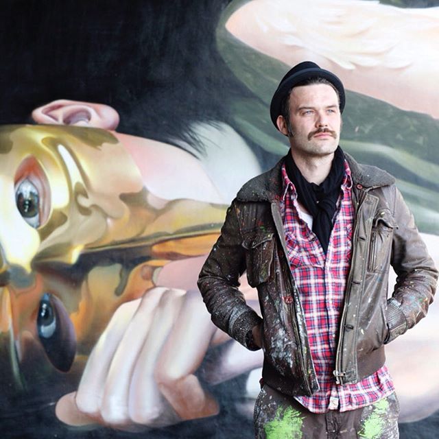 Man with a mustache poses in front of his large, surreal painting in a studio.