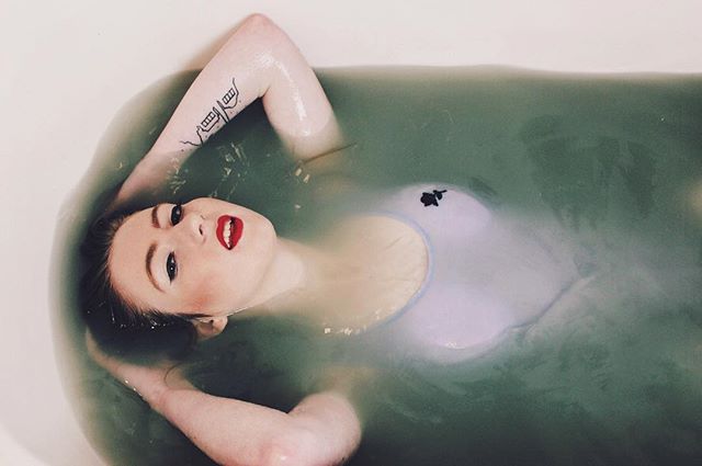 A woman with red lipstick relaxes in a tub of green water, showcasing an arm tattoo and a light-colored bathing suit.