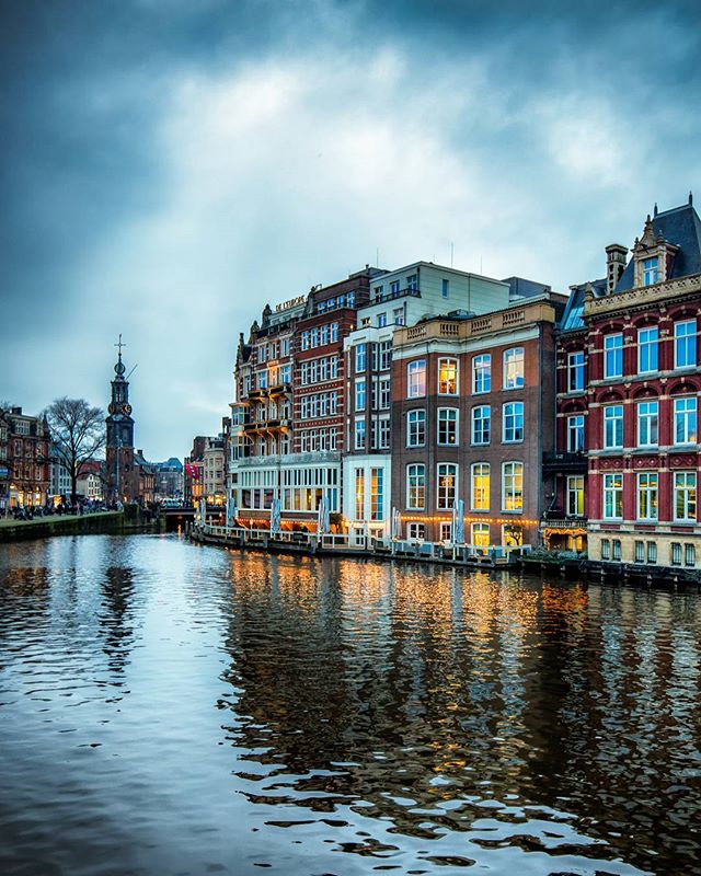 Scenic view of Amsterdam's canals and historic buildings under a cloudy sky, reflecting city lights in the water.