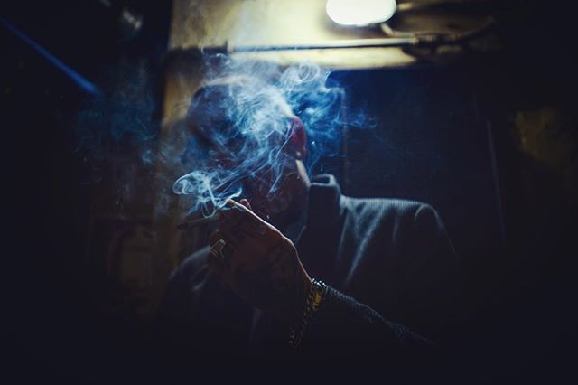 A man shrouded in smoke holds a cigarette in a moody, atmospheric indoor setting.