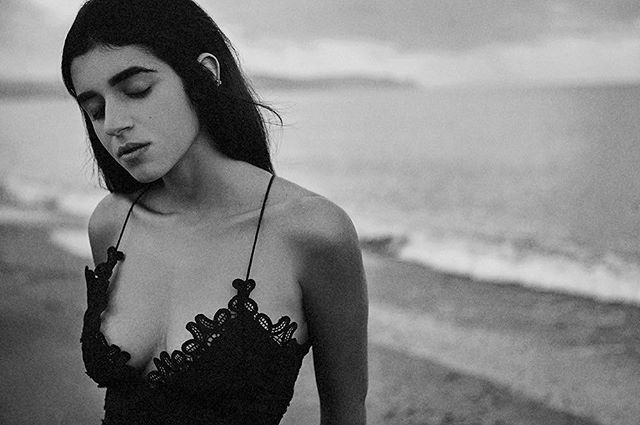 A woman in a black lace dress stands on a beach with a dreamy, serene mood in black and white.