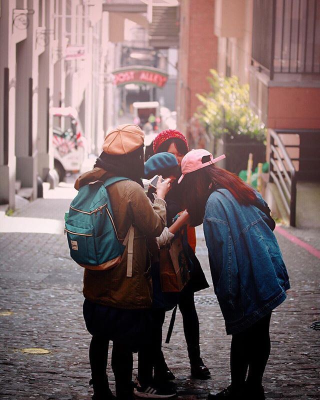 Four friends stand together on a cobblestone street in a city alley, looking at a phone and smiling.