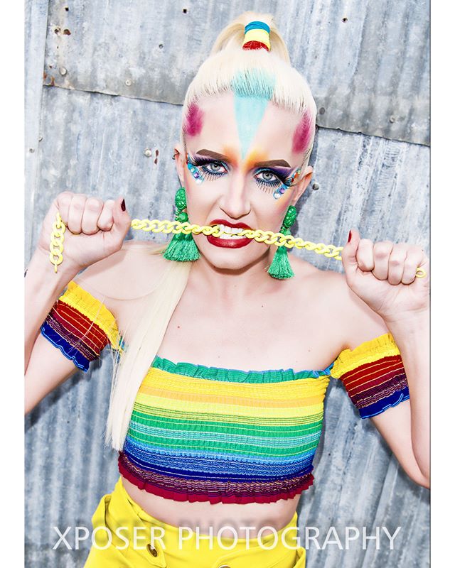 A woman with bright, rainbow makeup and a matching outfit bites a yellow chain in a playful editorial shot.