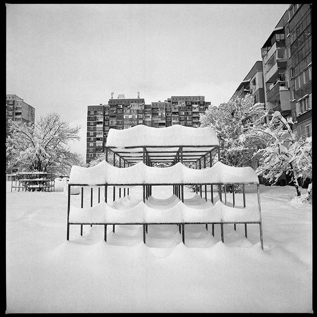 A black and white scene of snow covered playground equipment in an urban setting.