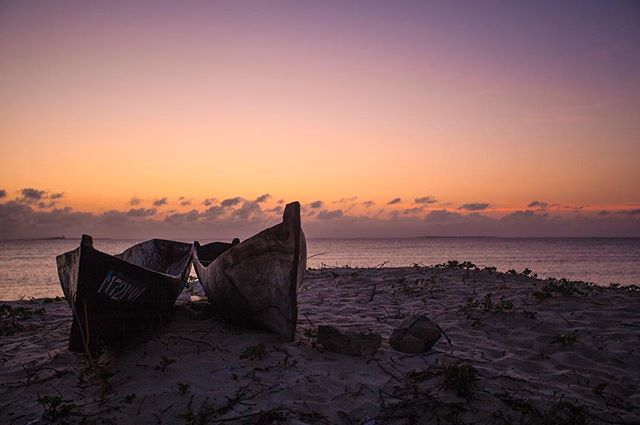 Two boats sit on a sandy beach as the sun sets over the ocean in a serene coastal scene.
