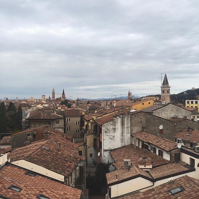 An aerial view shows the rooftops of a European city under a cloudy sky. The cityscape includes towers and historic buildings.