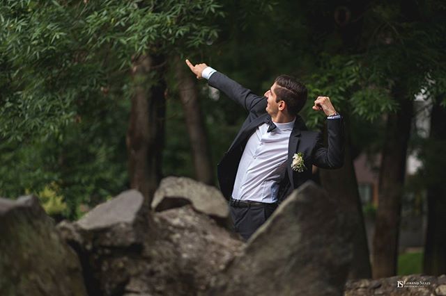 A groom in a suit playfully points towards something while celebrating outdoors in a park setting.