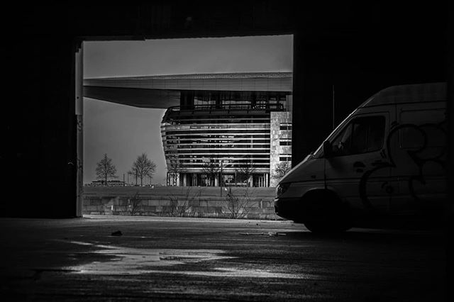 A grayscale image captures an urban scene with a modern building framed by an opening and a parked van.