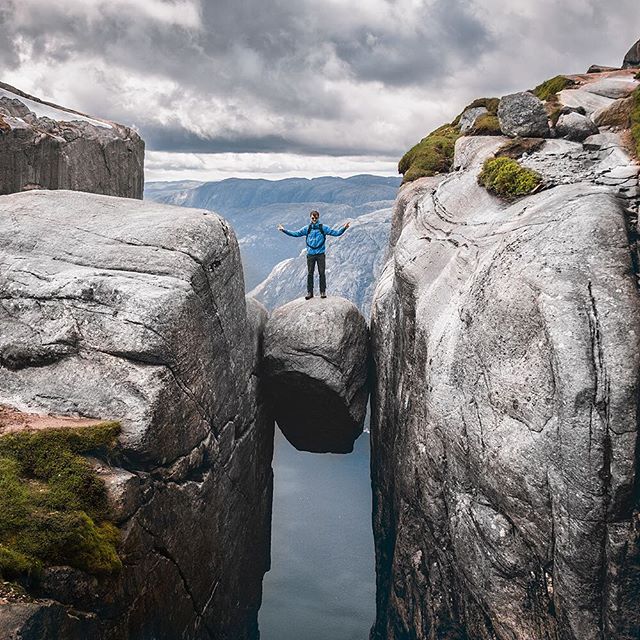 A man stands on Kjeragbolten, a rock wedged in a mountain crevice in Norway, against a dramatic sky and stunning landscape.