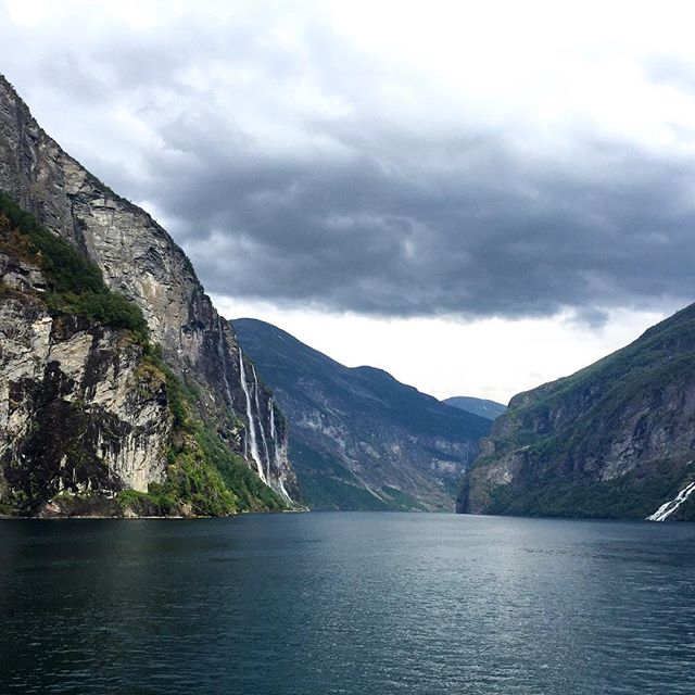 Dramatic fjord landscape with steep cliffs, cascading waterfalls, and calm waters under a cloudy sky.