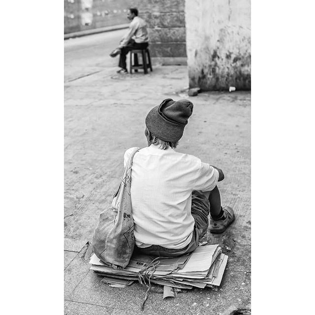 A man sits on cardboard in a black and white urban scene, with another man in the background.
