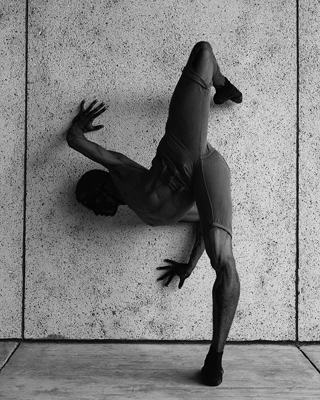 A male dancer performs a challenging pose against a concrete wall in a high-contrast black and white image.