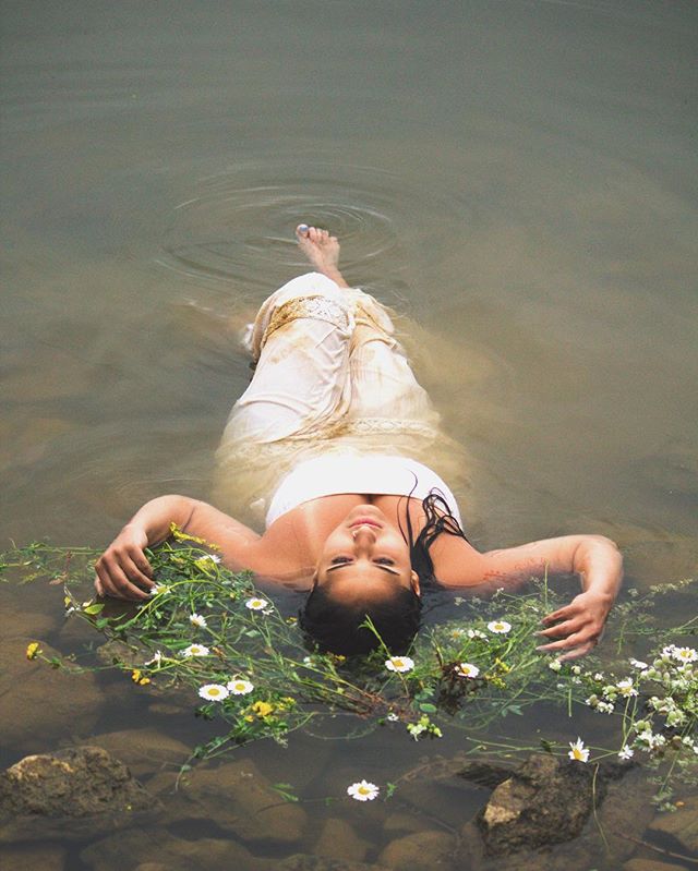A woman in a white dress floats serenely in a river surrounded by wildflowers.