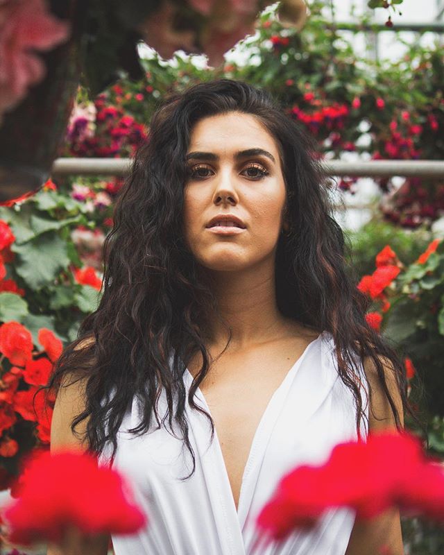 A woman with long curly hair poses in a garden setting surrounded by red flowers.